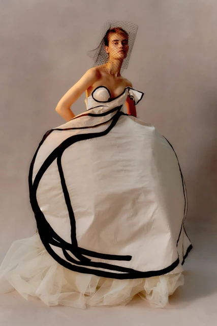 Woman in an ornate white strapless gown with black piping and black veil poses in a studio with a soft, neutral background.