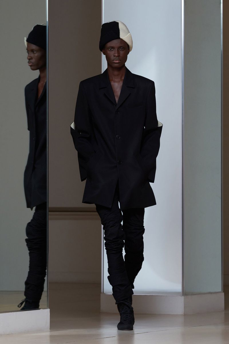 Runway fashion shot: A model in black jacket and pants walks by a mirror, wearing a color-blocked beanie in an indoor environment.