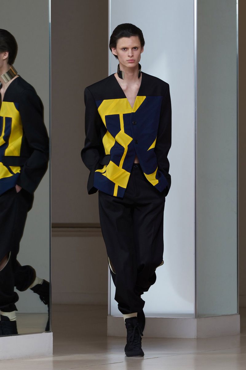 Model walks forward wearing a collarless black jacket with yellow and navy geometric patterning, black trousers, and a neck choker.