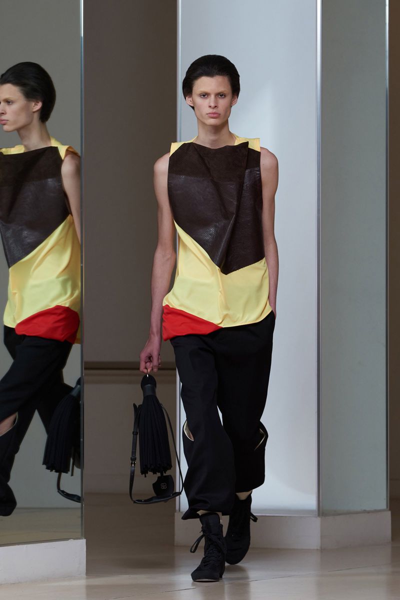 Runway shot of a fashion model showcasing a sleeveless, layered color-blocked top and black pants, holding a tassel purse.