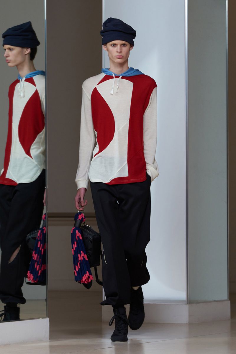 Runway fashion: a model presents a white and red long-sleeve top with black pants, accessorized with a blue beanie and patterned handbag.