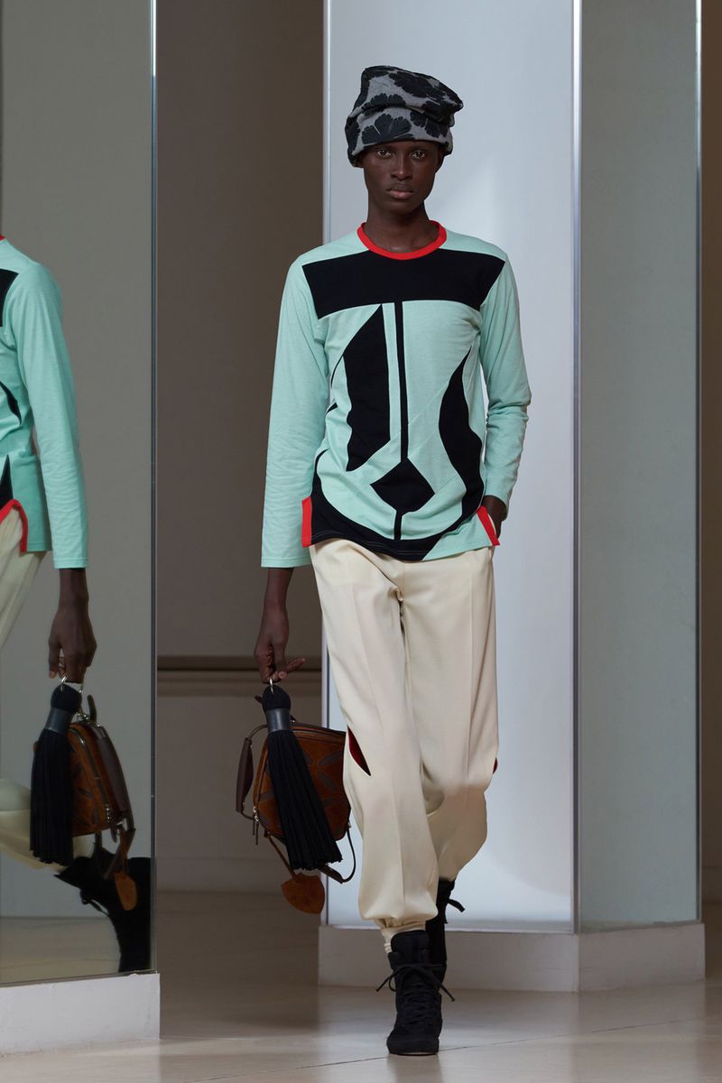 Model wearing patterned headwear, colour-blocked long-sleeve top and pale trousers holding a handbag
