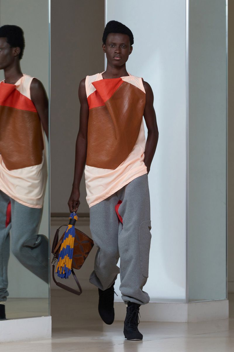 Runway fashion: A black male model showcases a unique top, sweatpants, and boots while carrying a leather handbag on a white runway.