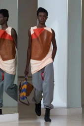 Black model walking wearing a sleeveless colorblock top, grey sweatpants, and holding a brown fringed handbag