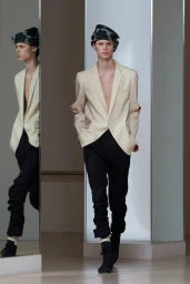 Model in cream blazer and dark trousers wearing a patterned hat standing before a mirror reflection