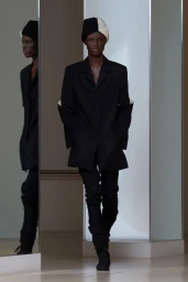 Model in black jacket and split black-and-white beanie posing in front of a mirror for Stefan Cooke