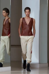 A model walking past mirrored pillars wearing a brown leather top and light trousers for Stefan Cooke F/W 24