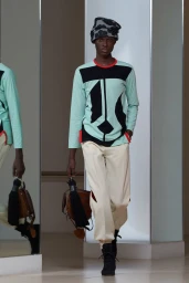 Model wearing patterned headwear, colour-blocked long-sleeve top and pale trousers holding a handbag