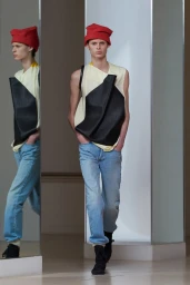 Model wearing a red beanie, sleeveless cream and black top, and light blue denim jeans walking past a mirrored column.