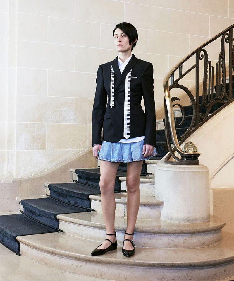A woman poses on marble stairs wearing a black blazer with piano key accents, a denim skirt, and black flats indoors.