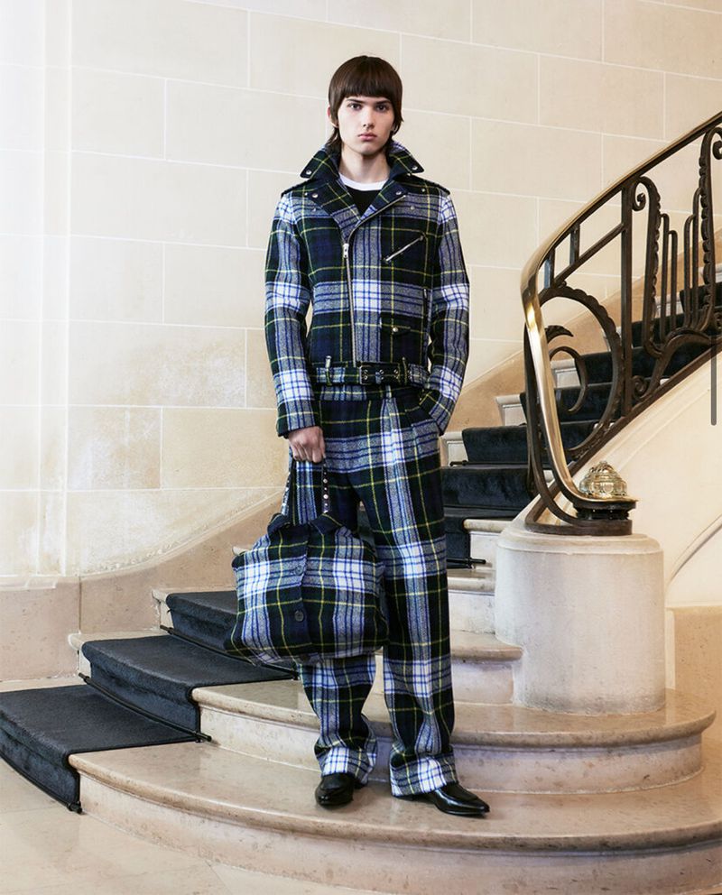 A male model wearing a plaid jumpsuit and carrying a matching bag stands on a marble staircase indoors, showcasing a unique fashion statement.