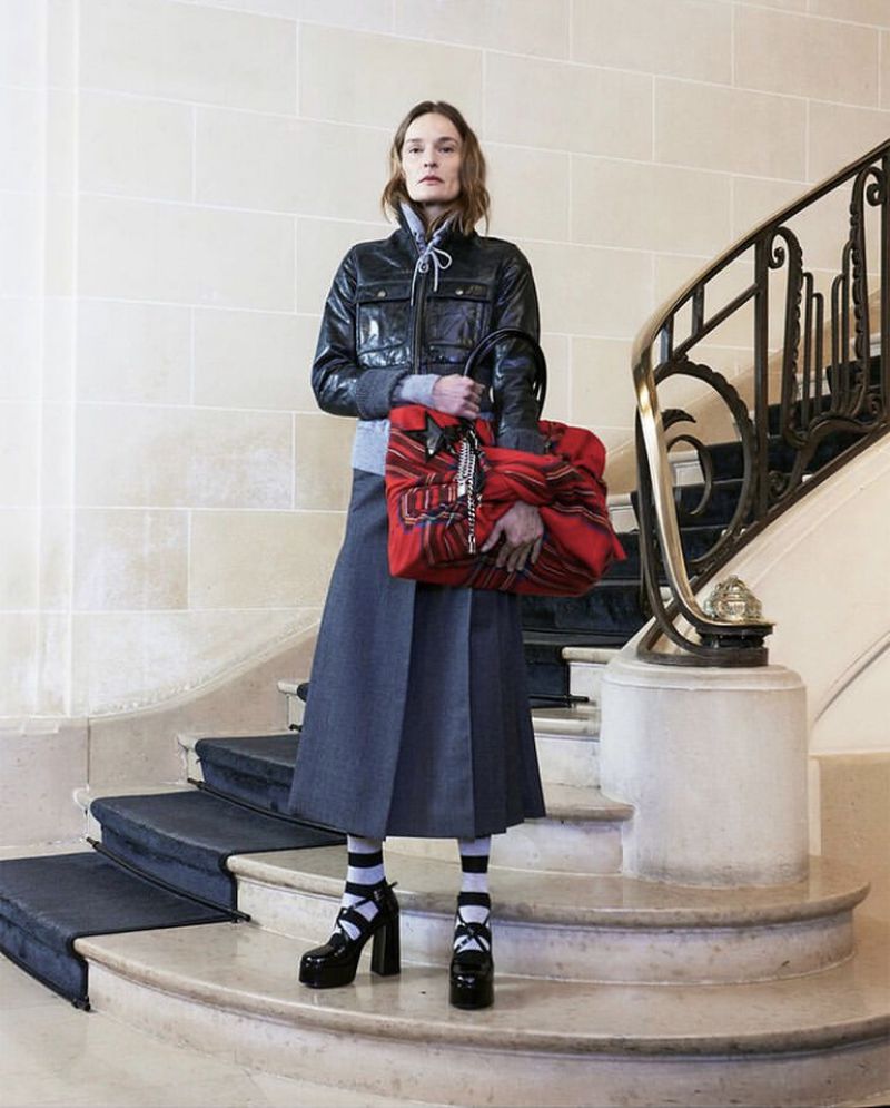Fashionable woman on marble staircase wearing a leather jacket, grey skirt, platform heels, and holding a patterned red tote bag.
