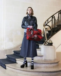 Model standing on stone stairs holding a large red patterned handbag wearing a black leather jacket and long grey skirt
