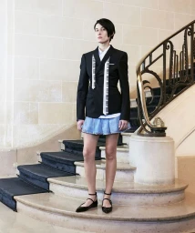 Model standing on stone staircase wearing a black blazer over white shirt and denim mini skirt for The Kooples F/W 24 Lookbook.