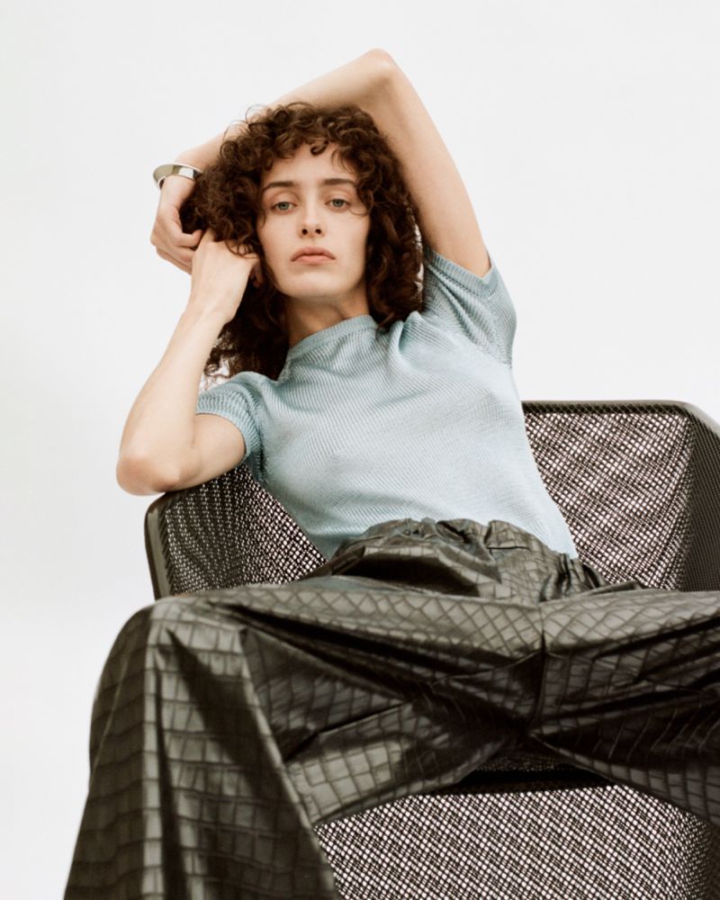 Model with curly hair seated in a dark chair wearing a light blue top and dark patterned trousers against white
