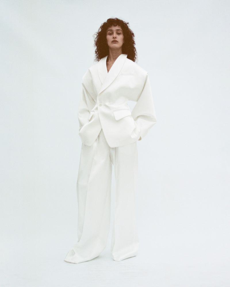 A woman with curly hair poses in an all-white oversized suit with a tie belt against a clean white backdrop in a studio environment.