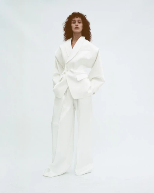 A woman with curly hair poses in an all-white oversized suit with a tie belt against a clean white backdrop in a studio environment.