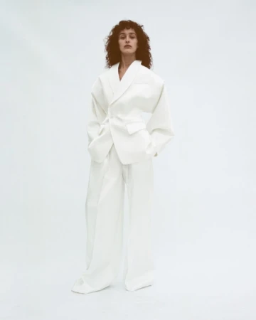 A woman with curly hair poses in an all-white oversized suit with a tie belt against a clean white backdrop in a studio environment.