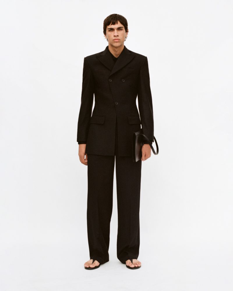 Man in black double-breasted suit stands in a photo studio, holding a black clutch. Minimalist fashion with a modern, elegant design.