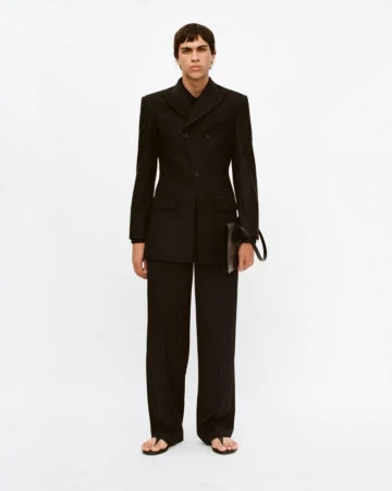 Man in black double-breasted suit stands in a photo studio, holding a black clutch. Minimalist fashion with a modern, elegant design.