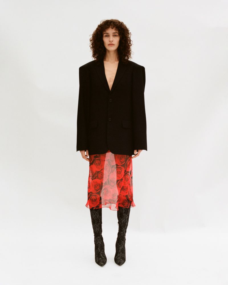 Woman in oversized black blazer, red rose-patterned dress, and black lace boots stands in a full shot against a white backdrop.