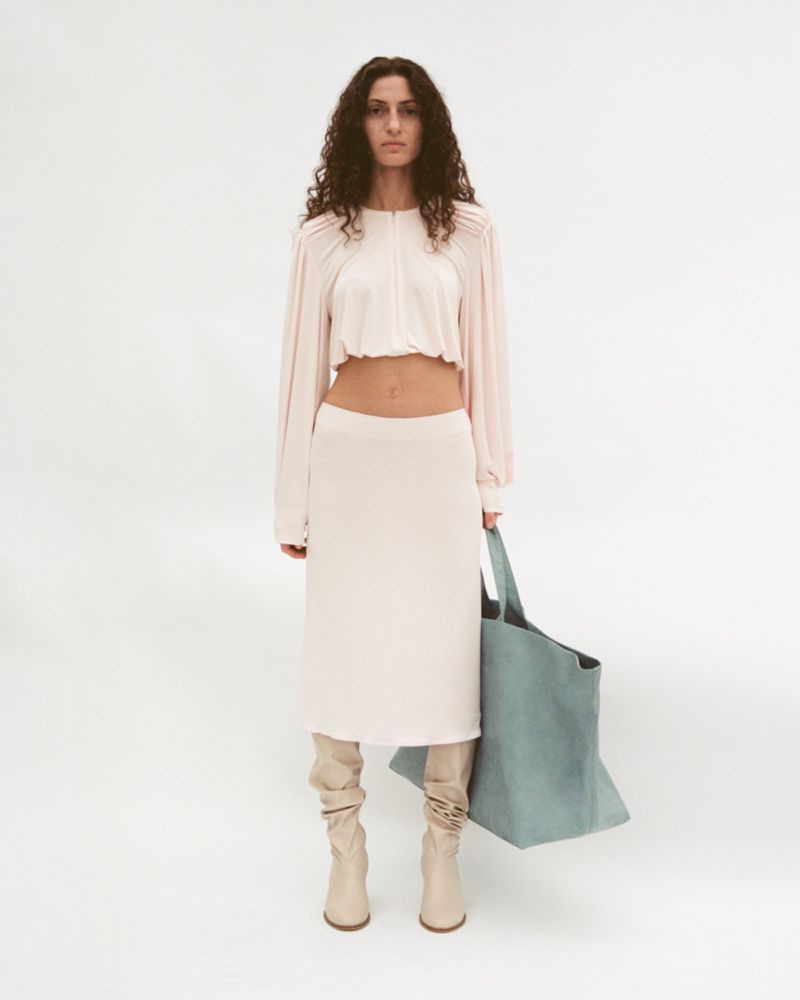 A woman in a pink top and skirt poses in a studio with a blue tote bag and beige boots against a white backdrop, fashion photography.