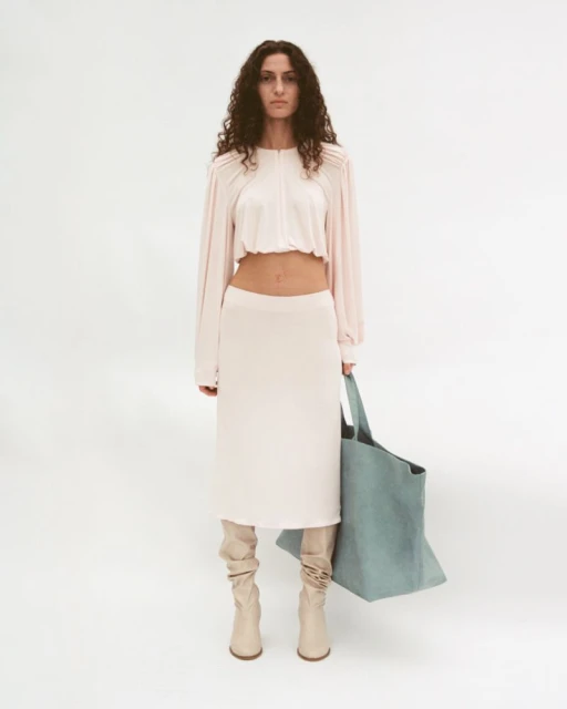A woman in a pink top and skirt poses in a studio with a blue tote bag and beige boots against a white backdrop, fashion photography.