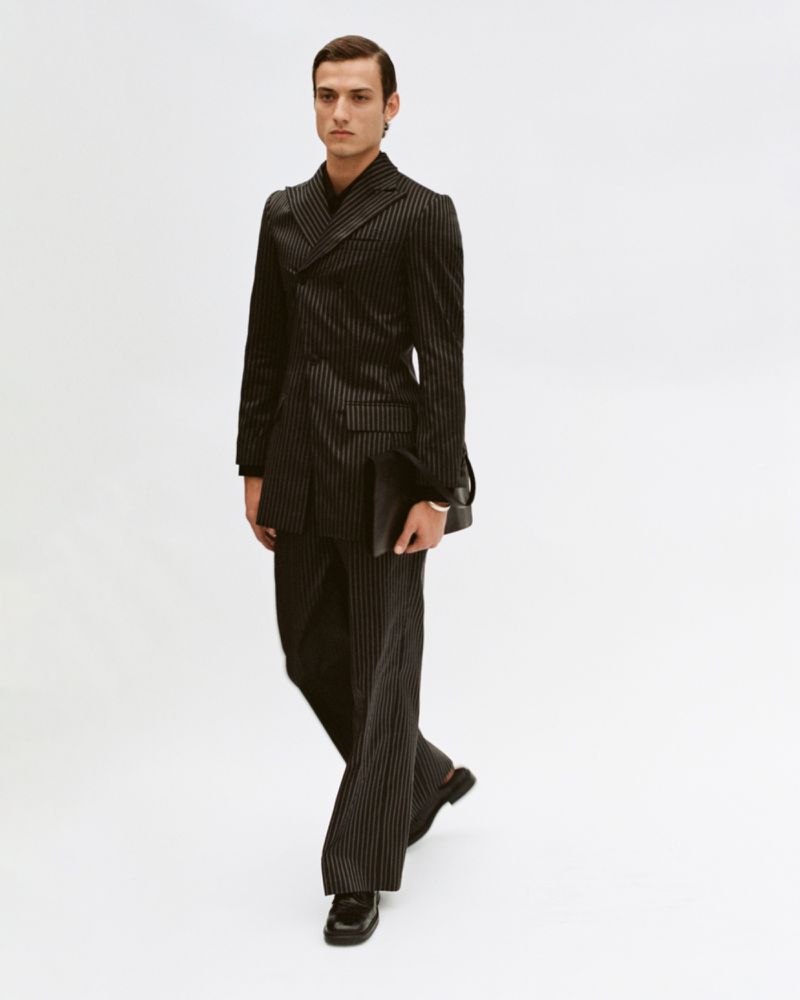 A man in a dark pinstripe suit walks toward the camera, holding a clutch, against a clean white backdrop in a studio setting.