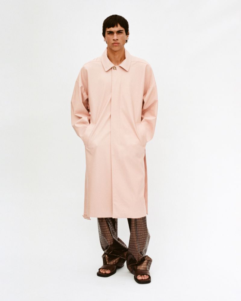 Full shot of a male fashion model posing in a studio wearing a long pink coat and patterned brown pants, showcasing current trends.
