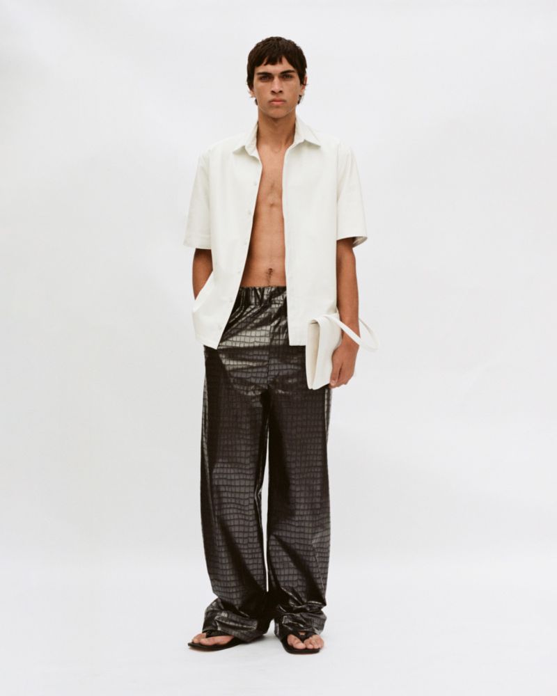Male model in an unbuttoned white shirt and dark patterned trousers holding a white clutch against a seamless background