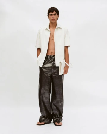 Male model in an unbuttoned white shirt and dark patterned trousers holding a white clutch against a seamless background