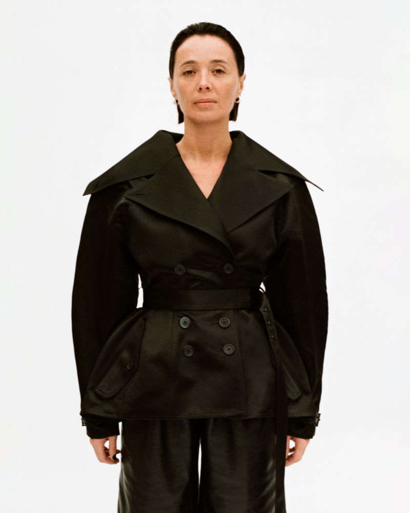 Adult female with dark hair wearing a black double-breasted short trench coat with a wide belt and black pants in a studio setting.