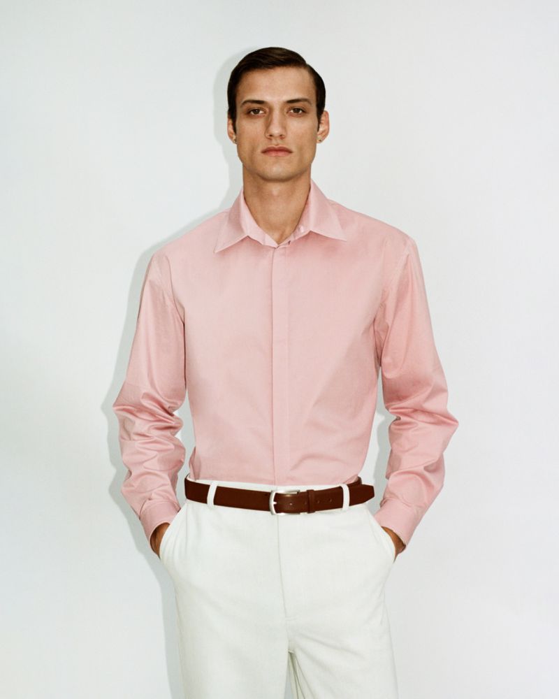 Model wearing a pink button-down shirt and white trousers with a brown belt against a plain white background