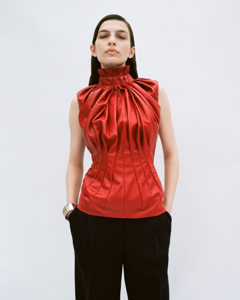 Model in a structured red high-neck top and black trousers against a neutral white seamless background.