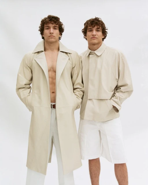Two adult males pose against a white background, one wearing a beige trench coat and white pants, the other a beige jacket and shorts.