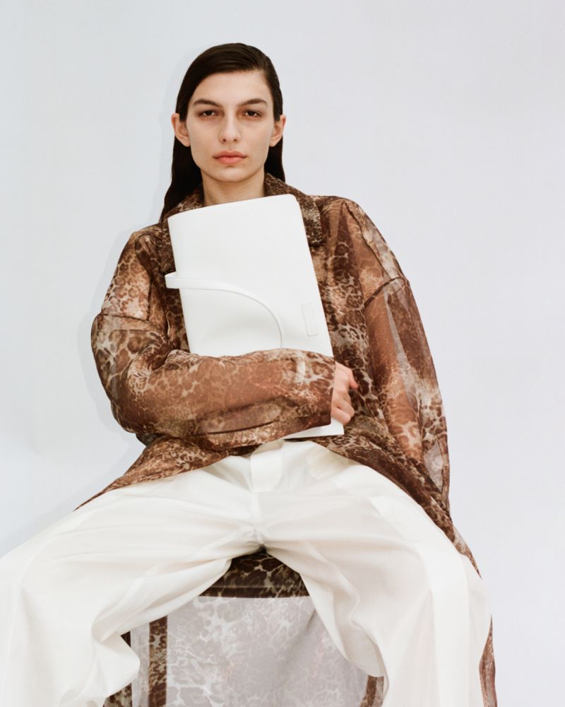 A woman in a sheer brown patterned robe holds a white bag, seated against a white background in a fashion-forward studio portrait.