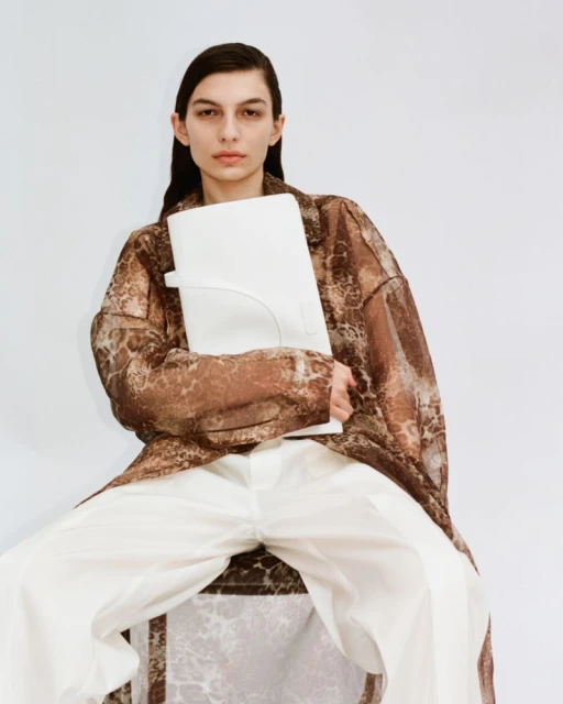 A woman in a sheer brown patterned robe holds a white bag, seated against a white background in a fashion-forward studio portrait.