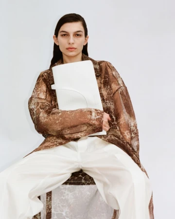 A woman in a sheer brown patterned robe holds a white bag, seated against a white background in a fashion-forward studio portrait.