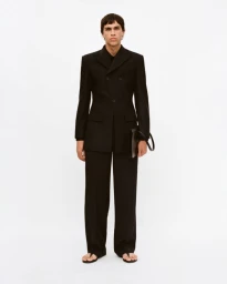 Model wearing a black tailored suit and holding a black handbag against a white background