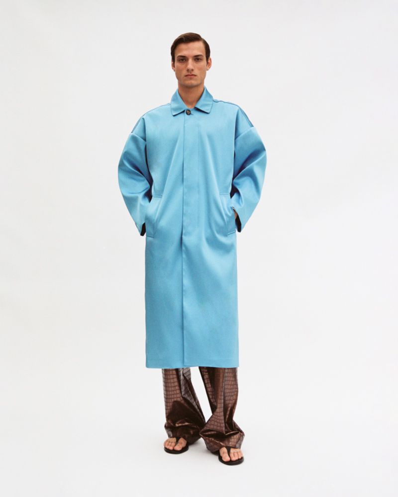 Model wearing a long blue coat with patterned brown trousers and sandals against a white background