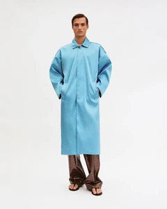 Model wearing a long blue coat with patterned brown trousers and sandals against a white background