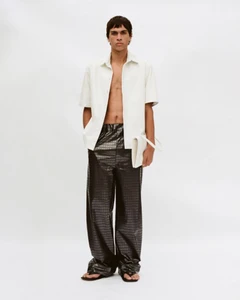 Male model in an unbuttoned white shirt and dark patterned trousers holding a white clutch against a seamless background