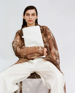Model with dark slicked-back hair wearing a patterned brown coat and white trousers holding a large white clutch