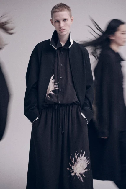 Stylish fashion shot of a young adult with short hair, wearing a black jacket and pants with light-colored floral designs. Studio lighting.