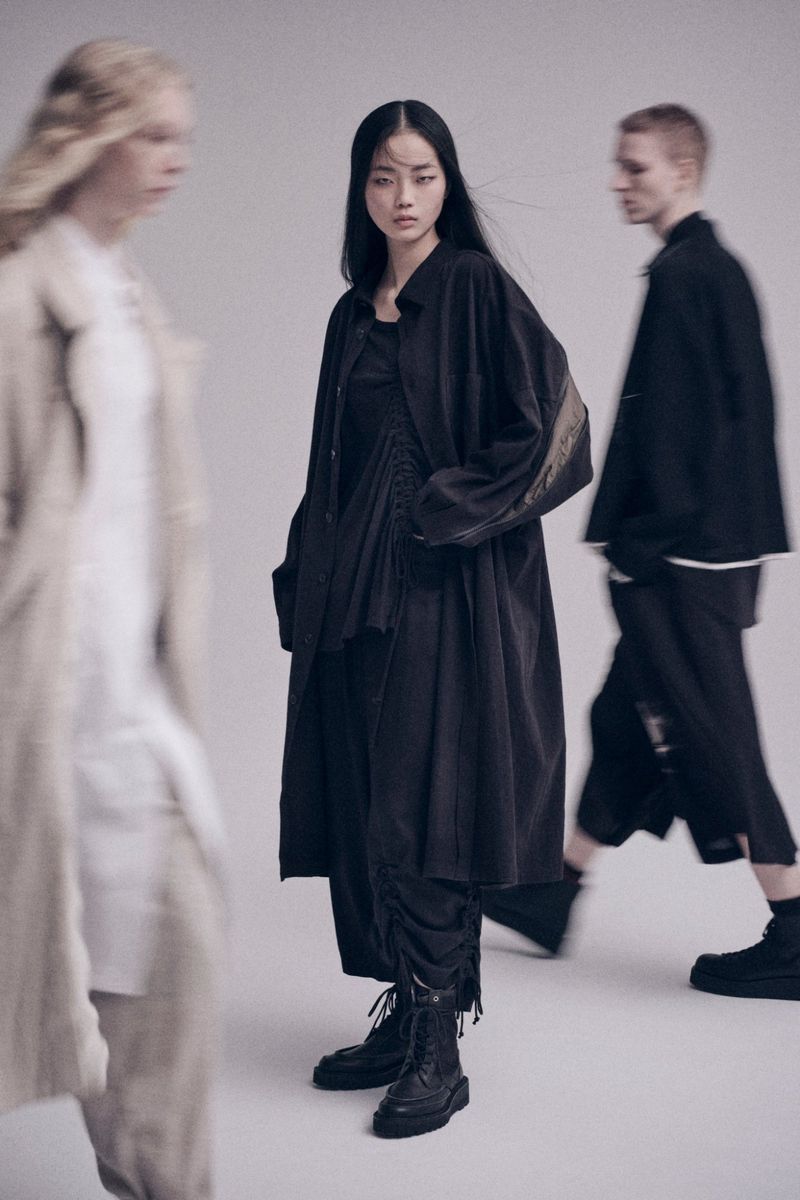 Three people wearing fashionable, dark, loose-fitting clothing pose in a photo studio. A woman stands centered in the frame.