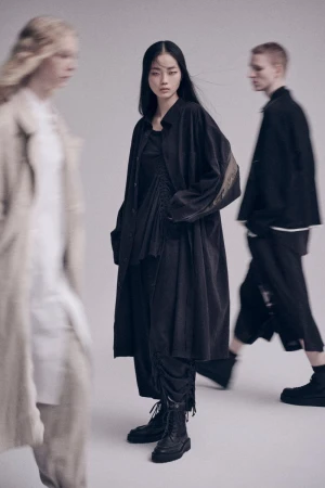 Three people wearing fashionable, dark, loose-fitting clothing pose in a photo studio. A woman stands centered in the frame.
