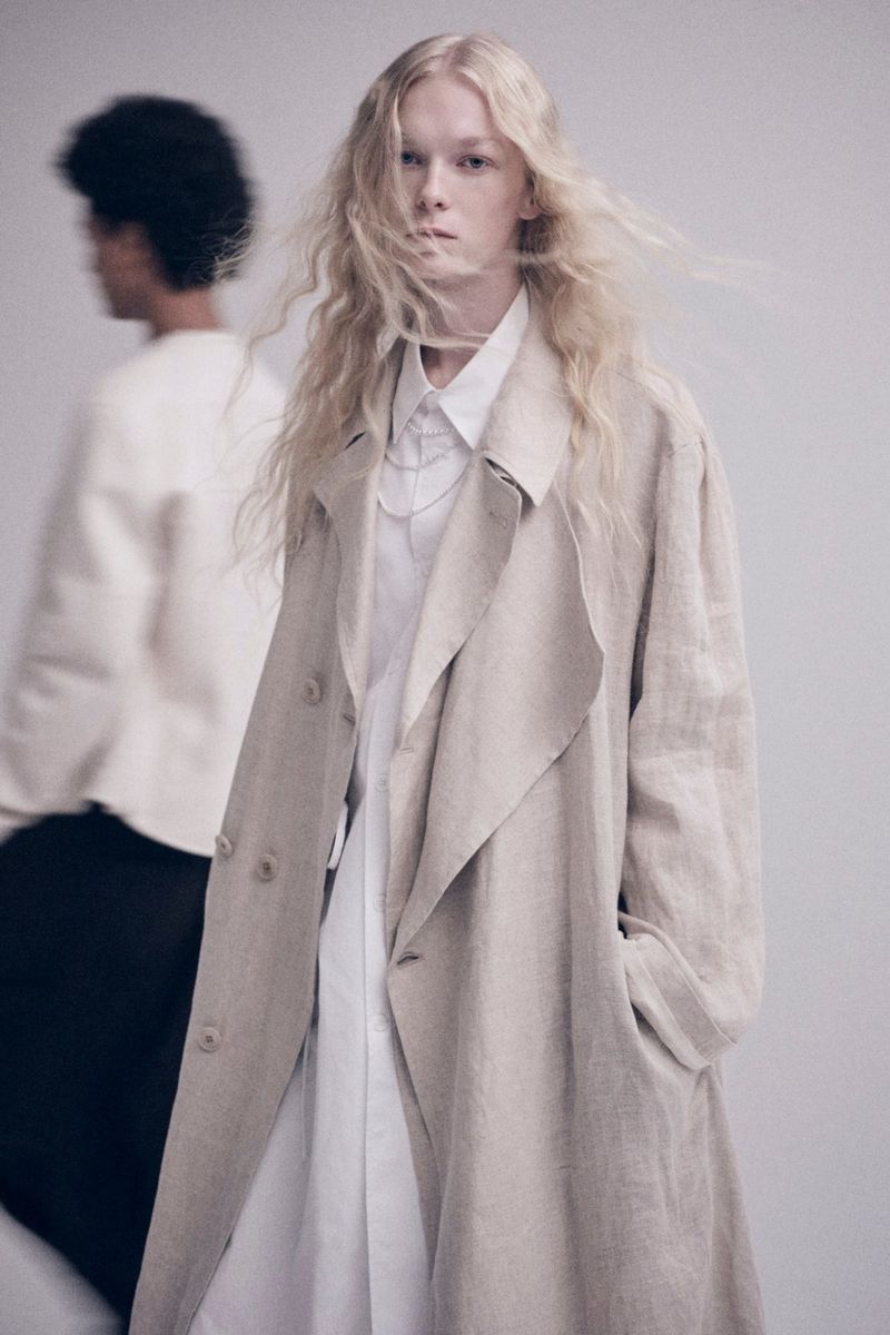 A fair-skinned woman with wavy blond hair poses indoors in a light beige trench coat and white shirt, with a blurry figure in the background.