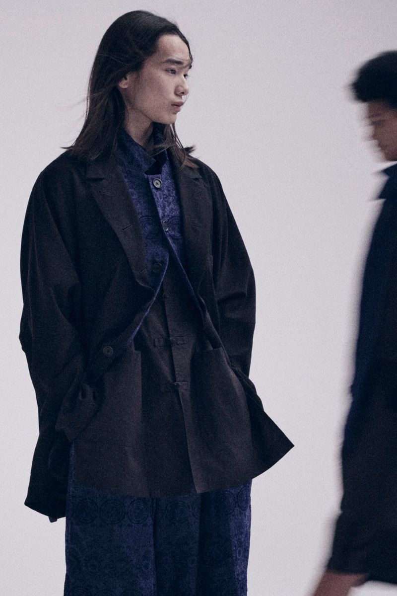 Two people model fashion in a studio setting. One person is in focus wearing a dark blazer and patterned shirt. Fashion editorial.