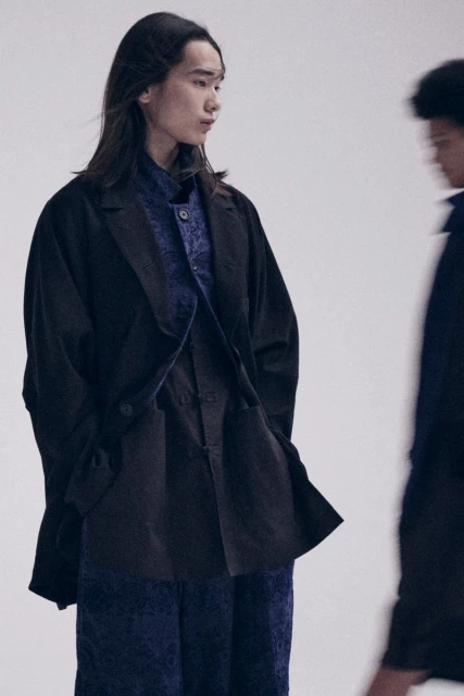 Two people model fashion in a studio setting. One person is in focus wearing a dark blazer and patterned shirt. Fashion editorial.