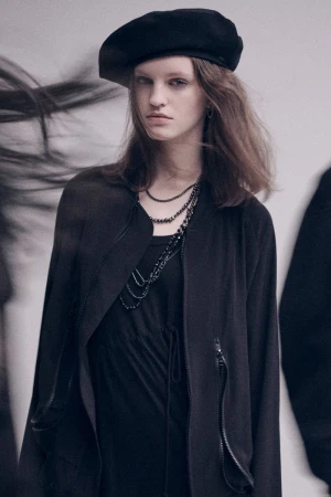 Model wearing a black beret and dark jacket with layered necklaces against a plain grey studio wall.